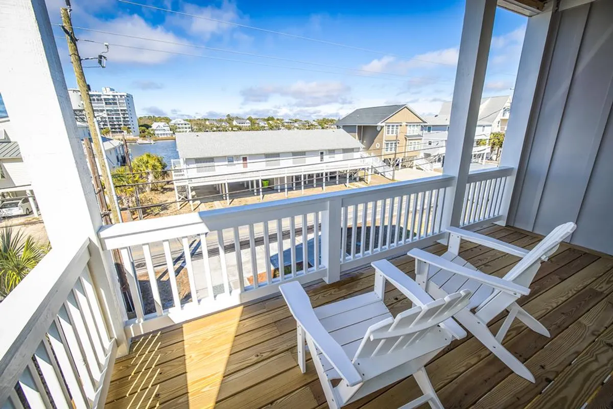 A serene exterior view features white Adirondack chairs on a wooden deck, with a vista of surrounding beach houses. The tranquil setting suggests places for family gatherings, peaceful sunset watching, and moments of solitude with the sound of the ocean nearby.