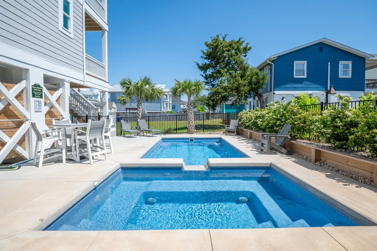 The image showcases a beautifully landscaped backyard with a sparkling private swimming pool surrounded by palm trees. There is an inviting outdoor seating area perfect for dining or lounging, with the relaxed vibe of Carolina Beach palpable under the warm glow of sunset light. The scene captures opportunities for swimming, sunbathing, and gathering with friends and family in an idyllic, tropical setting.