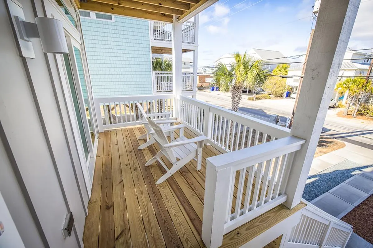 A serene wooden deck with white lounge chairs and palm trees, adjacent to blue house siding. The scene suggests relaxation, conversation, or taking in the view, with an inviting, open-air vibe.