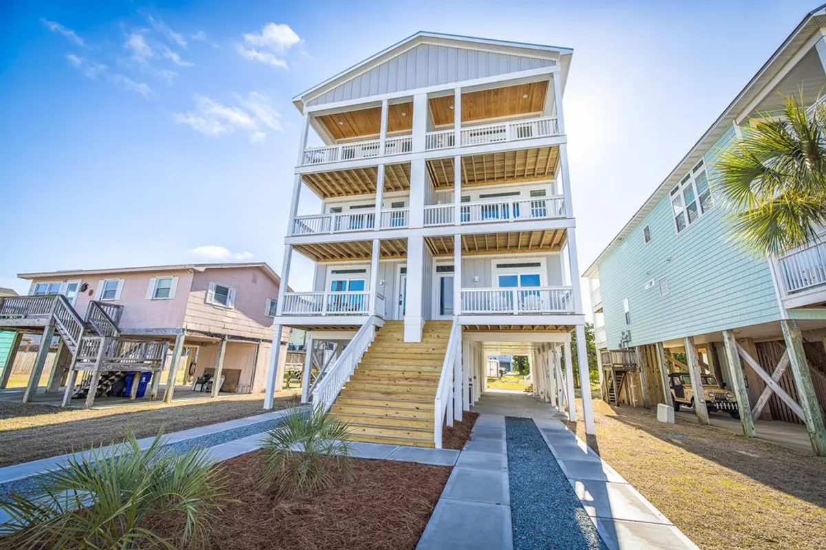 This image captures the exterior of a welcoming three-story beach house, featuring wooden stairs and inviting balconies. The sunset light enhances the warmth and hospitality of the space, while the outdoor areas hint at opportunities for relaxing mornings and evenings close to the shore.