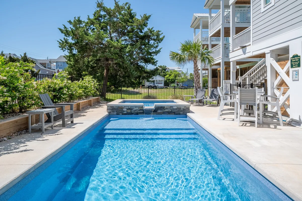 A vibrant outdoor scene showcasing a sparkling swimming pool bordered by a hot tub, surrounded by landscaped greenery and comfortable outdoor seating. The area is bathed in sunlight and gives off a welcoming, resort-like ambiance, ideal for relaxation and social gatherings.