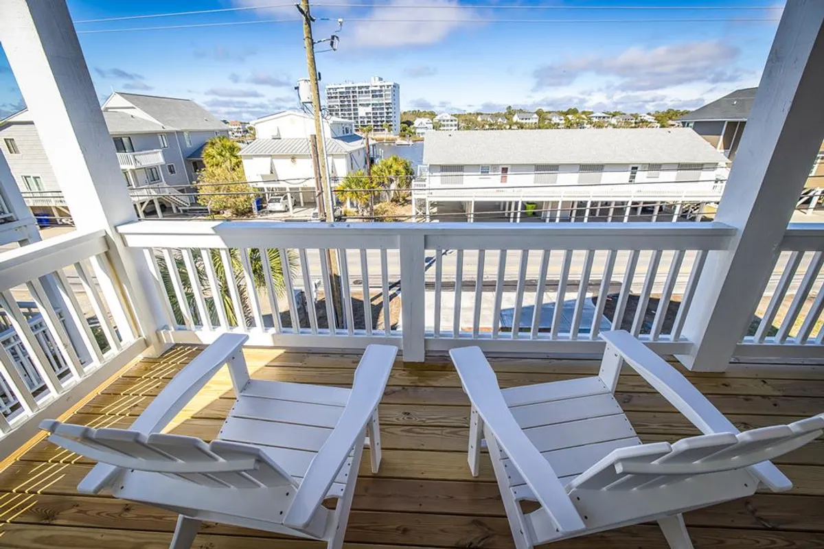 A tranquil balcony scene features wooden chairs facing a railing with a view over nearby homes and glimpses of the water. The setup is ideal for soaking in sunsets, enjoying morning light, or simply watching the world go by from a private vantage point.