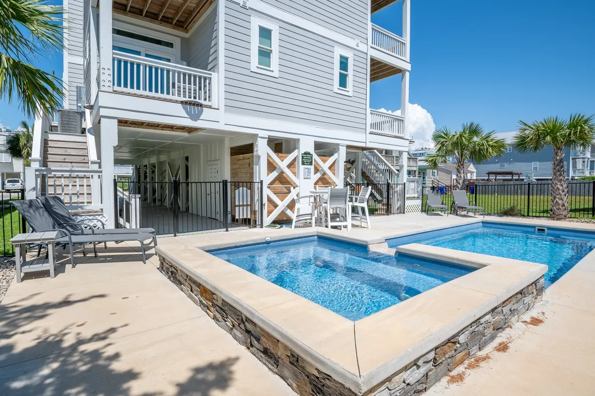 A sun-drenched poolside area featuring a swimming pool bordered by multiple lounge chairs and shaded by palm trees, with the exterior of the house visible in the background. The scene invites relaxation and socialization in a true vacation setting.