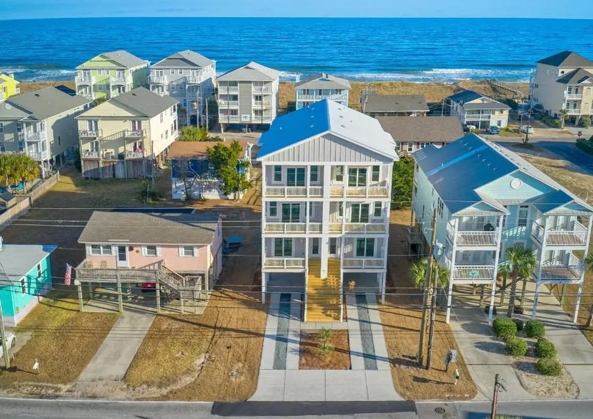 A striking view of a tall, modern house with palm trees lining a path, set against a backdrop of the sea. The foreground invites thoughts of gatherings and outdoor activities, while the ocean glimmers in the distance, conveying proximity to the beach.