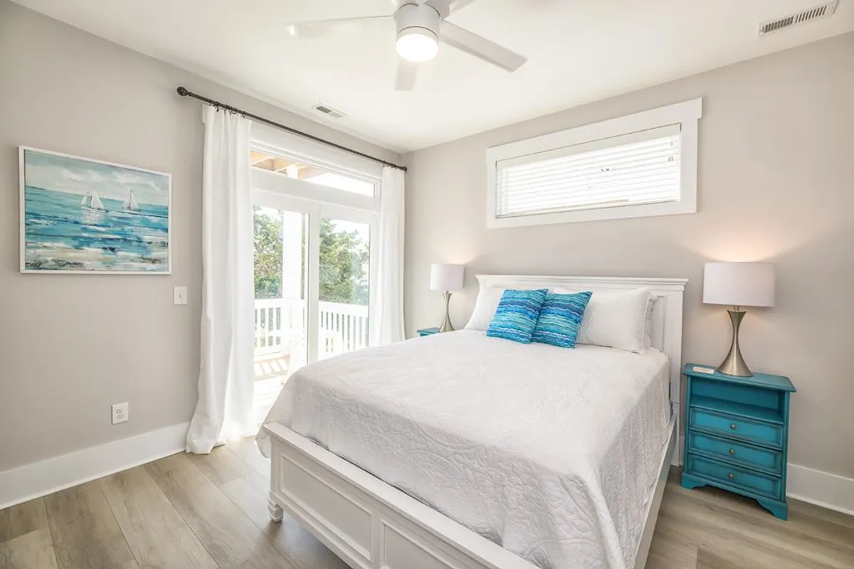 A tranquil bedroom scene featuring a cozy bed with blue decorative pillows, a painted bedside table, and art on the wall. The space is suffused with natural light, creating a restful atmosphere ideal for relaxation.